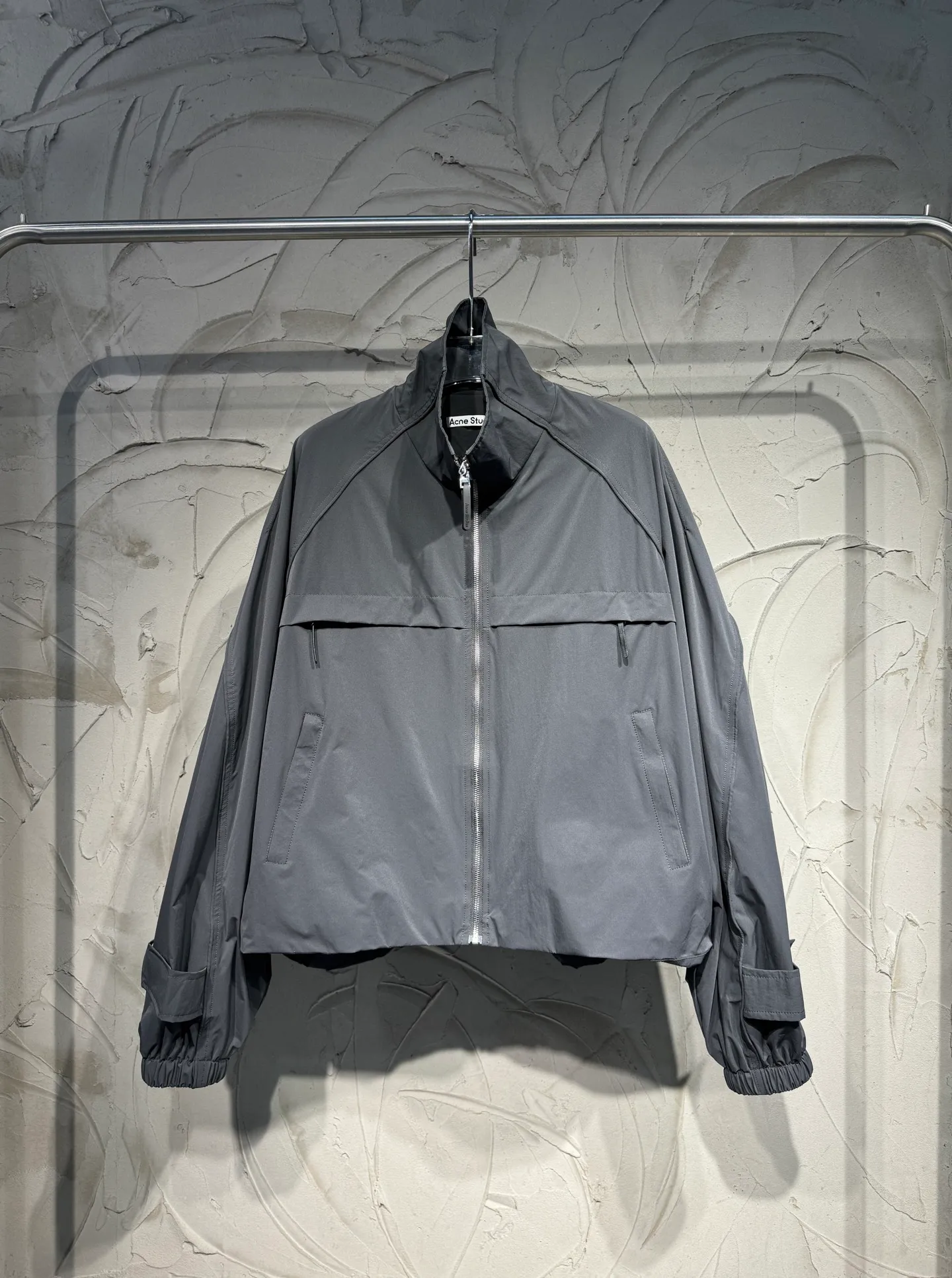 Acne Studios 26SS Large Lapel Short Jacket The fabric is made of 100% nylon (nylon/polyamide fiber) 75D high-density wrinkled nylon. The fabric has been permanently pressed and shaped by high temperature, and washed with high-pressure wrinkles at the factory to form its own irregular texture, permanently retaining the wrinkled texture, which will not disappear after washing. Matte matte coating removes the plastic glossy surface of nylon itself to create a low-key matte paper feel. Three-proof micro-coating, weak waterproof, anti-fouling, anti-static treatment, not easy to get dust. The interior is made of full-width mesh Breathable nylon lining, which reduces the stickiness of the nylon fabric to the body, improves air circulation, prevents sweating from sticking to the skin, and the zipper does not rub against the skin. The version adopts the raglan split topstitching process, and the front and rear panels are divided diagonally to expose even stitching. The three-dimensional dropped shoulders are loose and the fit is increased to increase the margin of movement. The back neckline is three-dimensional and high-density computer embroidered, and the embroidery lines are dense and not prickly. Customized all-metal matte nylon zipper, Genuine Leather embossed brand letters on the zipper leather tab, zipper tape with same color nylon fit to the body, double-locked reinforcement on both sides of the zipper to prevent pulling and tearing. Color: Gray Size: S M L